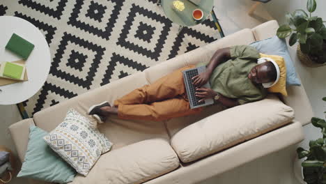 Hombre-Negro-Con-Auriculares-Descansando-En-El-Sofá-Y-Usando-Una-Laptop